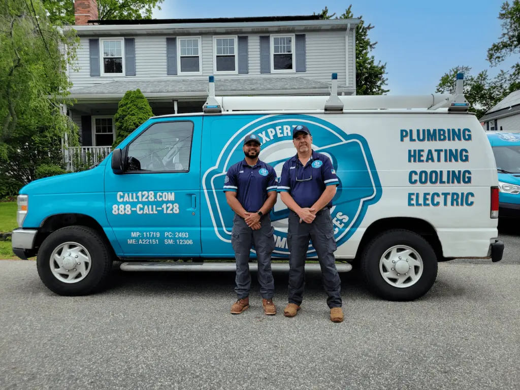 A plumbing, heating, cooling, and electric service van is parked on the street in front of a two-story suburban house with a manicured lawn and trees in the background.